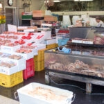 Hakodate Morning Market (函館朝市) – Fresh Seafood & Local Flavor in Hokkaido At Hakodate Morning Market, live crabs fill a tank and fresh seafood rests on ice in white trays atop yellow crates. A person stands behind the counter, with equipment and boxes visible in the bustling background.