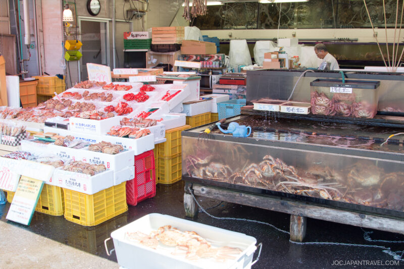 At Hakodate Morning Market, live crabs fill a tank and fresh seafood rests on ice in white trays atop yellow crates. A person stands behind the counter, with equipment and boxes visible in the bustling background.