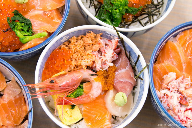 Bowls of Japanese chirashi sushi, inspired by the Hakodate Morning Market, topped with assorted seafood like salmon, shrimp, roe, and crab, plus fresh vegetables on rice with garnishes of wasabi and shredded seaweed.