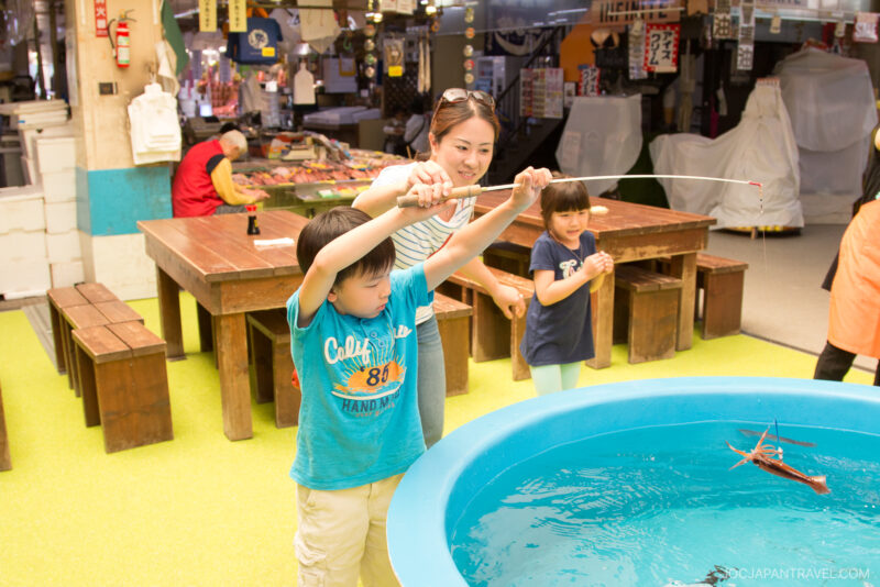 A woman helps a young boy use a fishing rod to catch a lobster from a blue tank, while a girl watches nearby inside the lively Hakodate Morning Market filled with tables and benches.