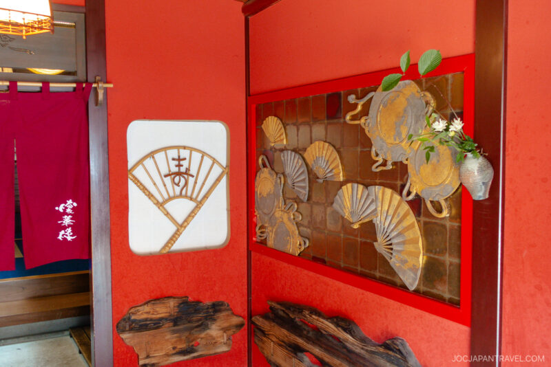 A vibrant red entryway at Higashi Chaya features decorative fans and crabs on the wall, a wooden accent piece, a small vase with flowers, and red noren curtains adorned with white Japanese characters.