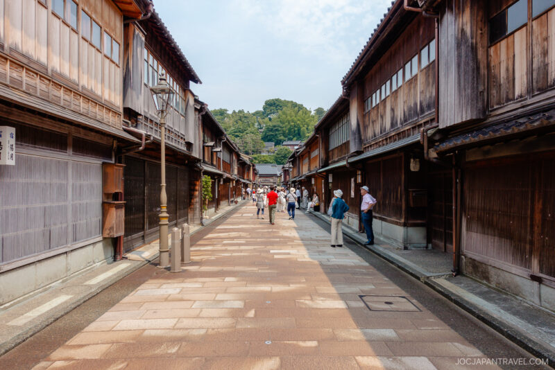People walk down a wide stone-paved street lined with traditional wooden buildings under a partly cloudy sky in Higashi Chaya, a historic Japanese district.