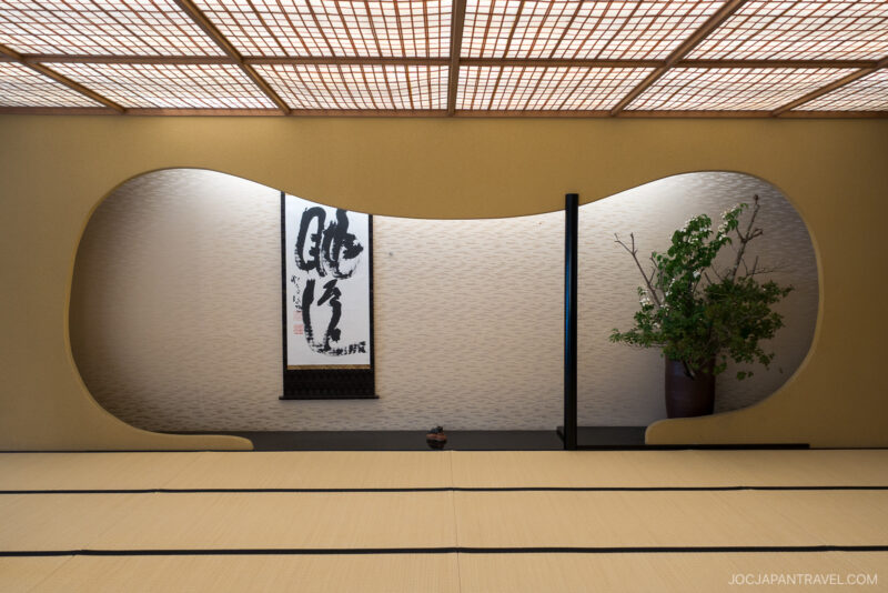A minimalist Japanese room at Honjin Hiranoya Kachoan Takayama features tatami mats, a curved wall opening, a hanging calligraphy scroll, a small pot, and a large vase with green branches, softly lit by a wooden lattice ceiling.