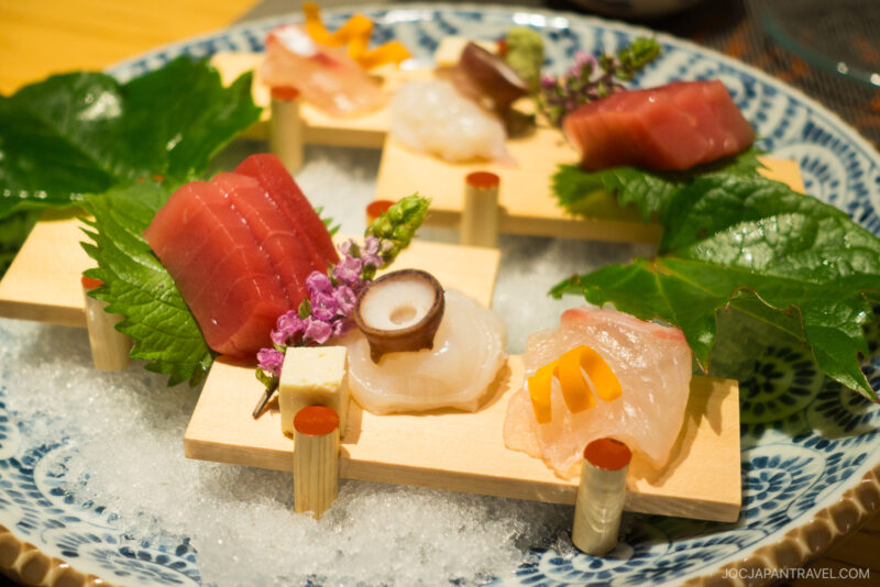 A plate of assorted sashimi, including tuna, octopus, and white fish, is artfully arranged on small wooden platforms over ice at Honjin Hiranoya Kachoan Takayama, garnished with shiso leaves and edible flowers.