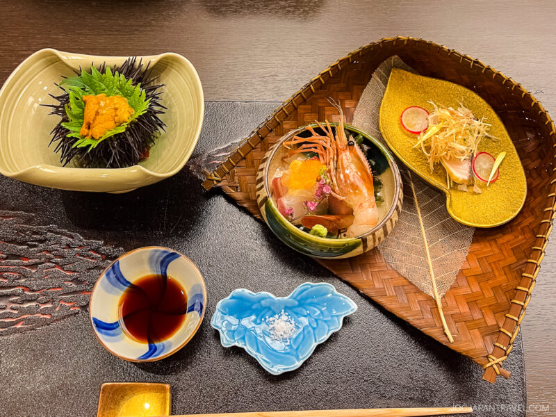 A Japanese meal with sashimi, sea urchin in its shell, a radish salad, soy sauce, and sea salt in a blue dish—all beautifully arranged on a woven tray atop a dark table at Hyakurakusou.