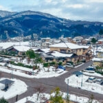 A snowy Japanese village, featuring houses, parked cars, and trees, nestled among snow-covered mountains beneath a cloudy sky.