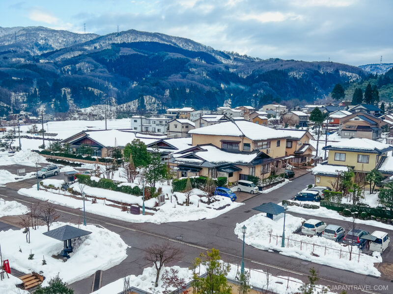 A snowy Japanese village, featuring houses, parked cars, and trees, nestled among snow-covered mountains beneath a cloudy sky.