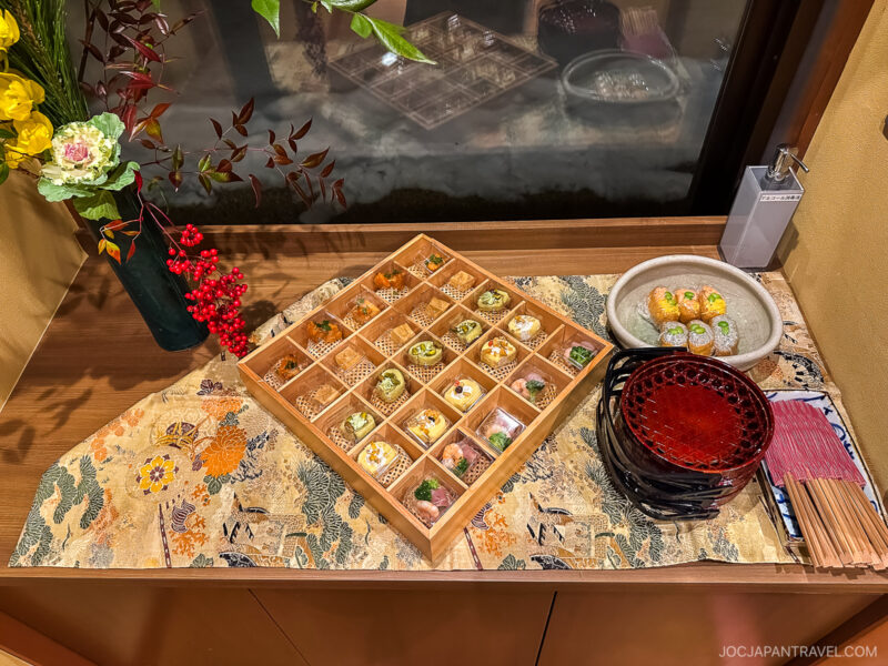 A wooden tray with assorted Japanese desserts from Hyakurakusou, sushi rolls on a plate, and stacked red lacquer bowls sit on a decorative table runner beside flowers and red berries, near a window.