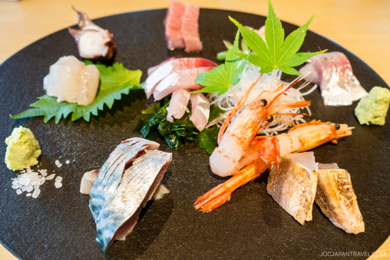 At Izakaya Dai Toyama, a black plate with assorted sashimi—shrimp, fish, and octopus—is garnished with a green maple leaf, shiso leaf, wasabi, and salt, all artfully arranged on a wooden table.