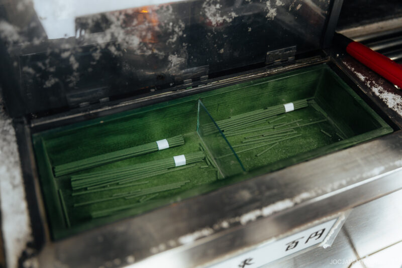 A close-up of a green tray containing thin, green sticks with white labels inside a glass-covered box, likely used for omikuji, reflecting Japanese Shrine and Temple Etiquette in an Asian setting.