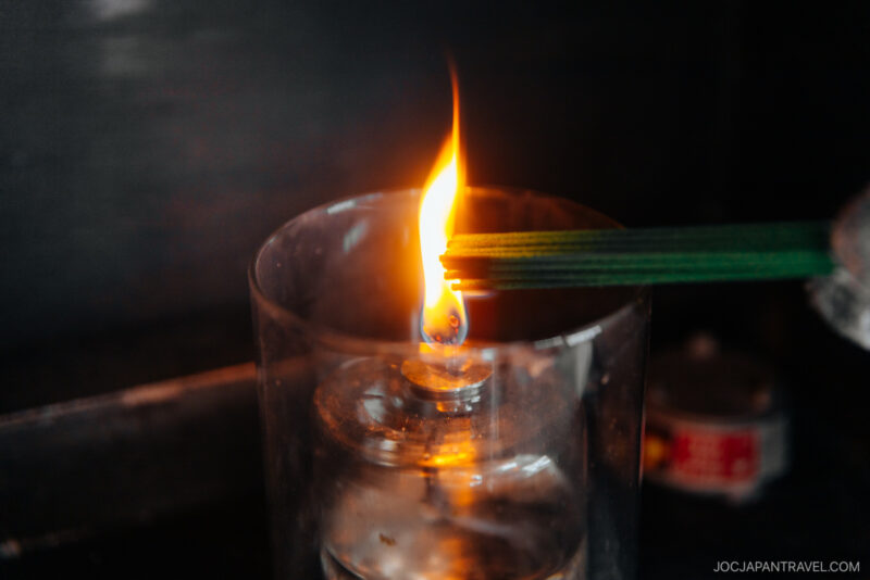 A lit alcohol lamp with a visible orange flame and several green incense sticks held over the flame, evoking Japanese Shrine and Temple Etiquette, set against a dark background.