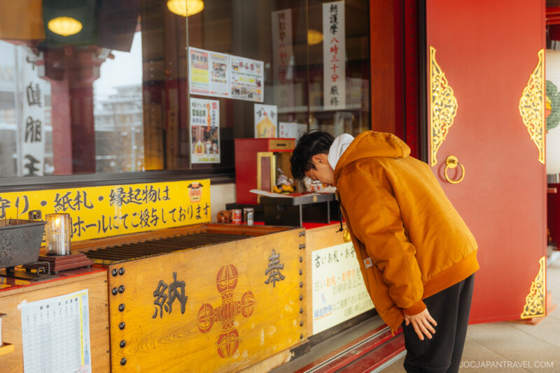 A person in an orange jacket bows at a wooden offertory box in front of a colorful shrine, practicing Japanese Shrine and Temple Etiquette, with signs and ornate red and gold details visible around the entrance.
