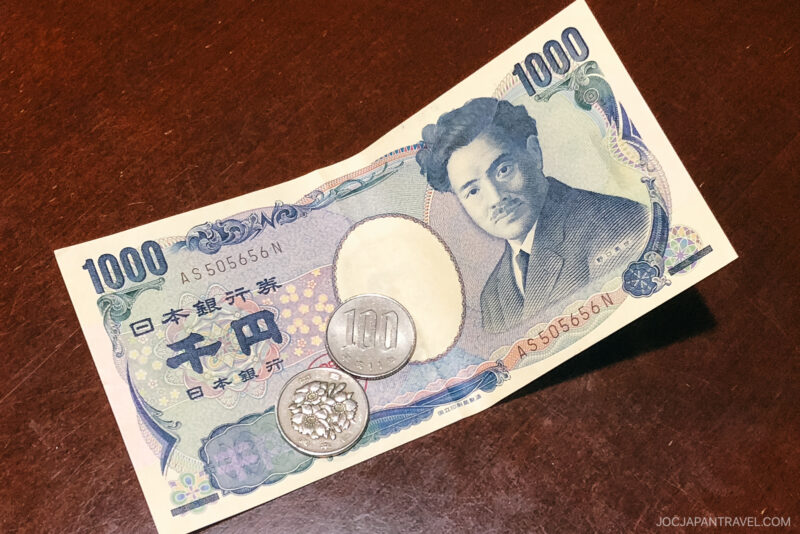 A 1000 yen Japanese banknote and two coins, one silver 100 yen coin and one 10 yen bronze coin, placed on a dark wooden surface—an everyday sight featured in any Japanese Etiquette Guide.