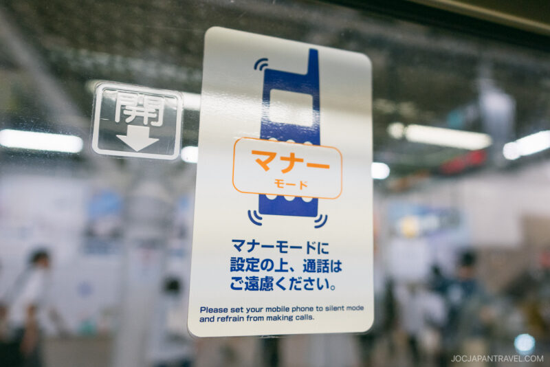 A sign on a glass window in a public place, styled like a Japanese Etiquette Guide, instructs people in both Japanese and English to set mobile phones to silent mode and avoid calls. Blurry figures and objects can be seen in the background.