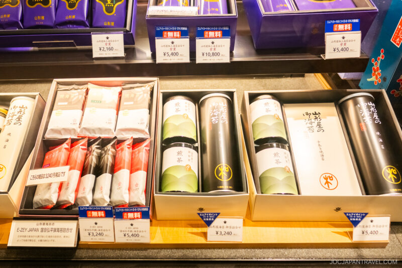 Gift boxes of Japanese tea and sweets are displayed on a store shelf, each with price tags in Japanese yen. The elegant packaging reflects traditional colors and design, making them perfect gifts for those interested in a Japanese Etiquette Guide.