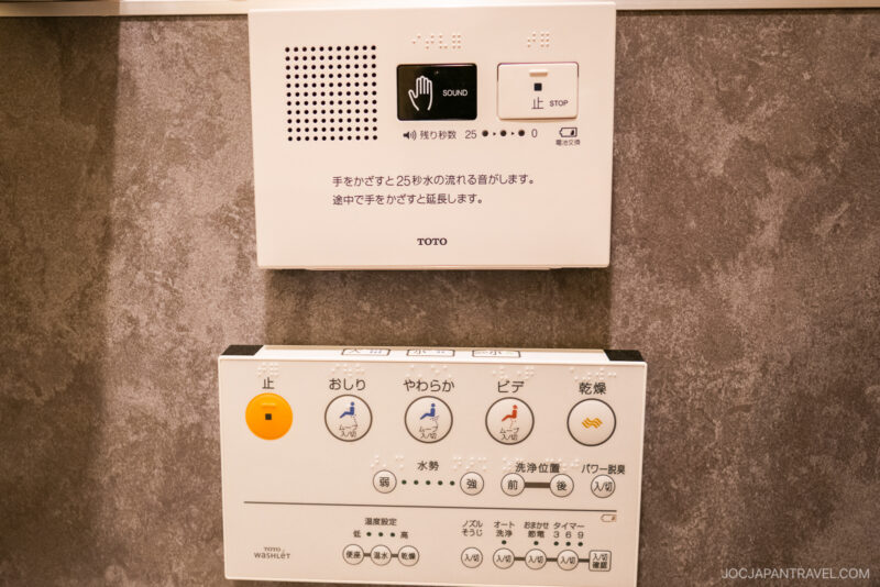 Japanese toilet control panel with various buttons and labels for functions like bidet, water spray strength, drying, and stop, mounted on a wall—an example often featured in a Japanese Etiquette Guide. The panel includes symbols and Japanese text instructions.