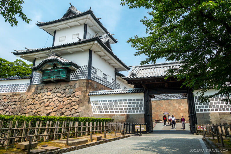 A traditional Japanese castle entrance with white walls, tiled roofs, and a large wooden gate welcomes visitors to Kanazawa Castle Park. Several people walk through the gate as trees frame the scene under a clear sky.