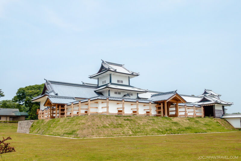 Kanazawa Castle Park features a traditional Japanese castle with white walls and blue-tiled roofs, perched on a grass-covered stone foundation beneath a clear sky.
