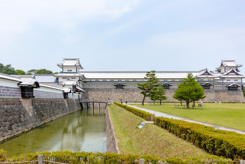 Kanazawa Castle Park features a traditional Japanese castle with white walls and tiled roofs, set behind a stone moat and surrounded by green lawns, trees, and neatly trimmed hedges under a clear sky.