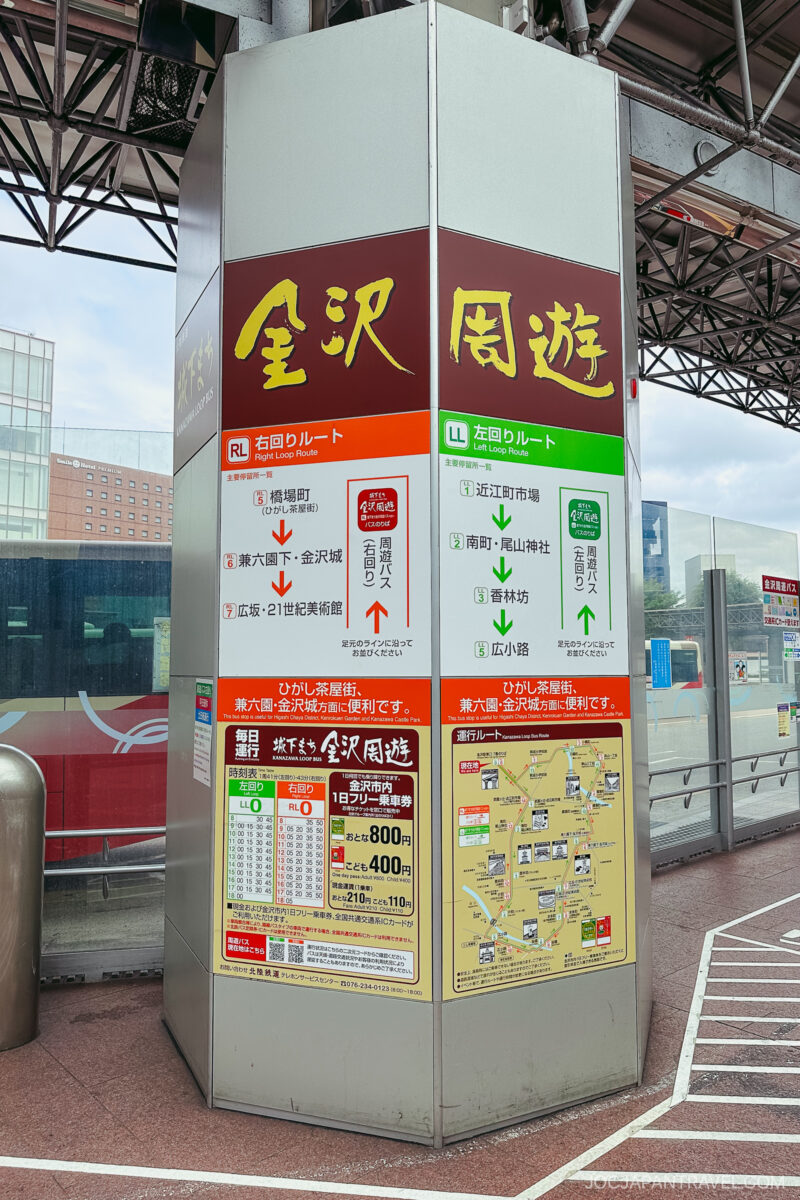 A sign at a bus station in Kanazawa, Japan, displays route information, schedules, maps, and fares for the Kanzawa Loop Bus. The sign features red, yellow, and green sections with Japanese and English text.