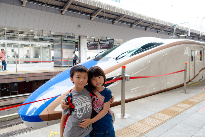 Two children stand arm in arm at a train station platform in front of a sleek, modern Shinkansen Kagayaki. The train is white with blue and brown accents, while the covered roof and signs add to the bustling atmosphere.