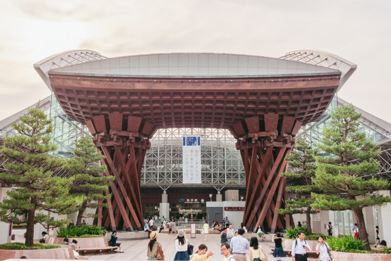 The large wooden gate with unique curved supports marks the entrance of Kanazawa Station’s modern glass building, surrounded by trees and people walking in front. The sky is clear and bright.