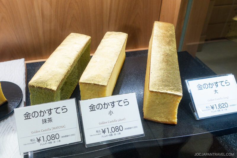 Three rectangular Gold Leaf Kasutera Cakes are displayed behind glass, each covered in gold leaf and labeled with their names and prices in Japanese and English. The cakes come in various sizes and flavors.