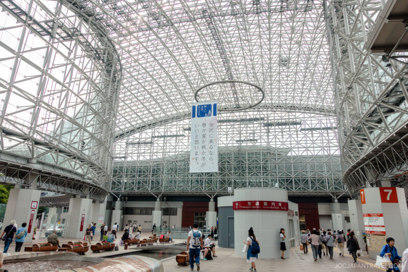 A spacious indoor plaza at Kanazawa Station features a glass and steel arched roof, busy with people walking and sitting. Signs in Japanese hang from the ceiling, and numbered red pillars mark different areas.