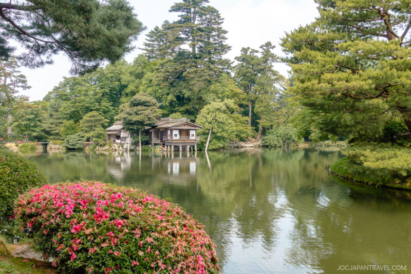A tranquil Japanese garden scene inspired by Kenrokuen Garden, with a wooden teahouse by a serene pond, surrounded by lush green trees and blooming pink azaleas under a partly cloudy sky.