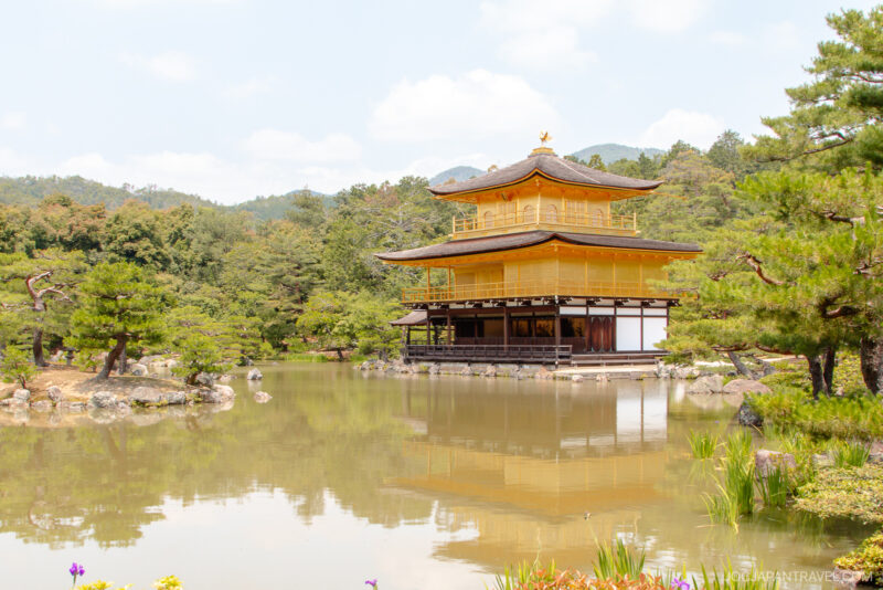 A golden pavilion stands beside a tranquil pond, surrounded by lush green trees and gardens under a partly cloudy sky at Kinkaku-ji Temple in Kyoto, Japan.