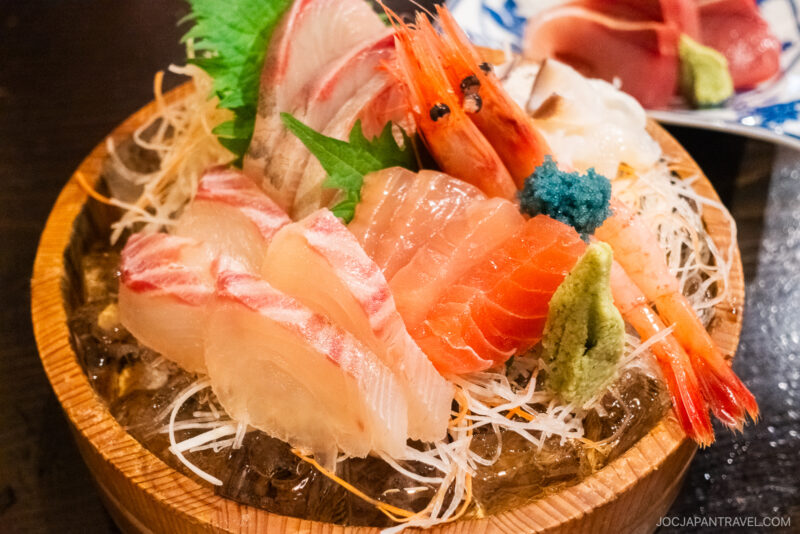 At Kuroya Izakaya Kanazawa, a wooden bowl filled with assorted sashimi—salmon, tuna, shrimp, and garnishes like wasabi, shredded daikon, and shiso leaf—is elegantly served on a bed of ice.