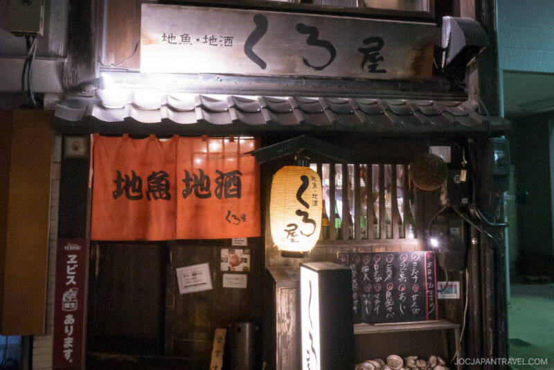 Kuroya Izakaya Kanazawa is a traditional Japanese izakaya with a wooden exterior, red and brown signs in Japanese, a hanging paper lantern, and a chalkboard menu outside the entrance at night.