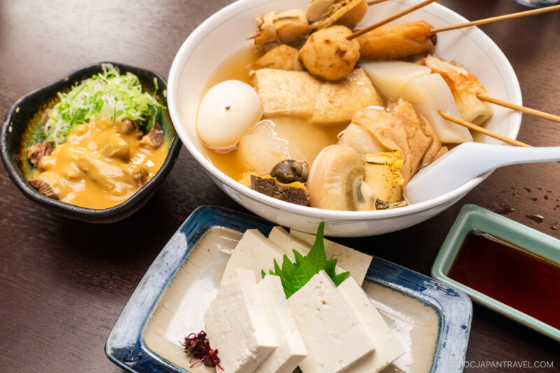 A bowl of Kuroyuri Oden Kanazawa with assorted skewered ingredients, a dish of fresh tofu slices with garnish, a small bowl of miso sauce with green onions, and a soy sauce dish on a wooden table.