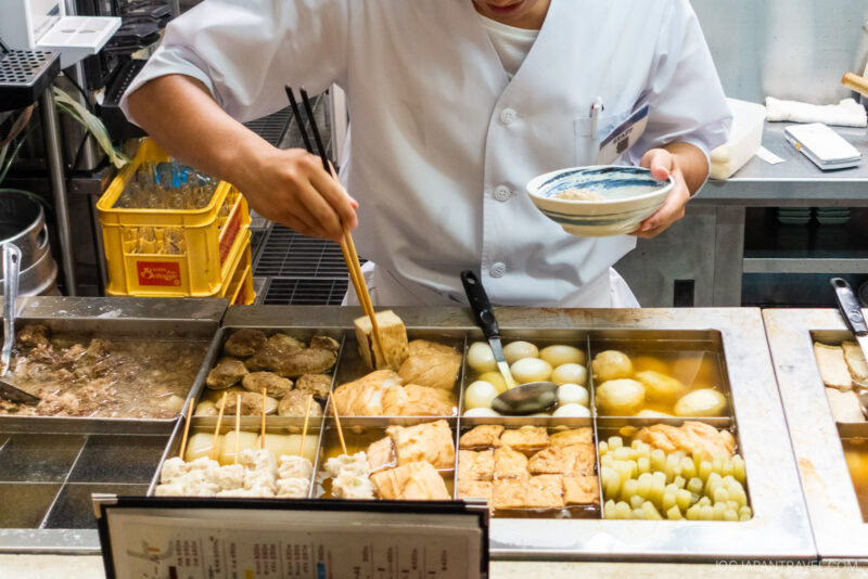 At Kuroyuri Oden Kanazawa, a person in a white chef’s coat serves Japanese oden from a steaming counter filled with simmering eggs, tofu, and vegetables, using chopsticks and a ladle.