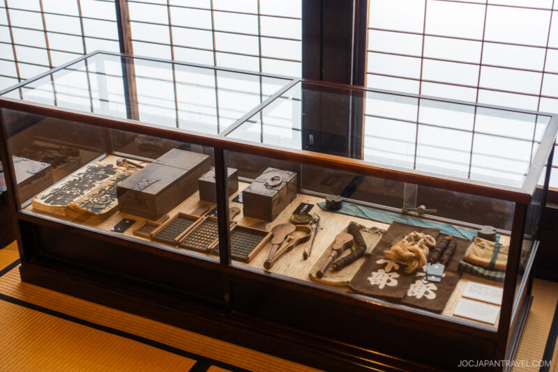 A glass display case in Kusakabe Heritage House contains traditional Japanese tools, wooden objects, books, abacuses, and cloth items—all arranged neatly on a tatami mat floor in a well-lit room with shoji screens.