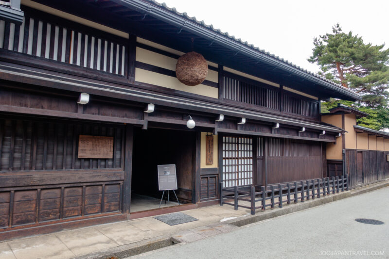 Traditional Japanese wooden building with dark timber exterior, sliding doors, and a large round brown sugidama above the entrance—this serene Kusakabe Heritage House sits on a quiet street lined with trees in the background.