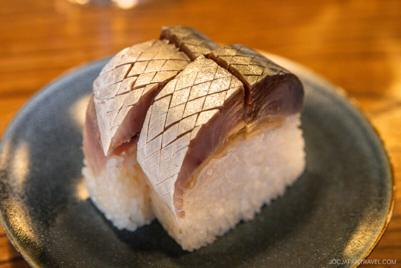 A close-up of two pieces of pressed sushi topped with shiny, silver-skinned mackerel on a round, dark plate. This Kyoto what to eat specialty features mackerel with a delicate crisscross pattern cut into the surface.