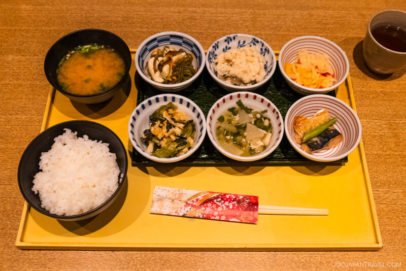A Japanese meal set on a yellow tray with white rice, miso soup, six small dishes of vegetables, tofu, pickles, tea, and decorated chopsticks—perfect for those wondering in Kyoto what to eat for an authentic experience.
