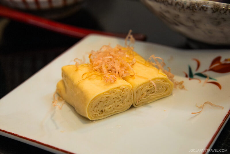 Two neatly rolled pieces of Japanese yuba (tofu skin), a must-try for anyone wondering Kyoto what to eat, are topped with bonito flakes and served on a decorative white plate with a red floral design.