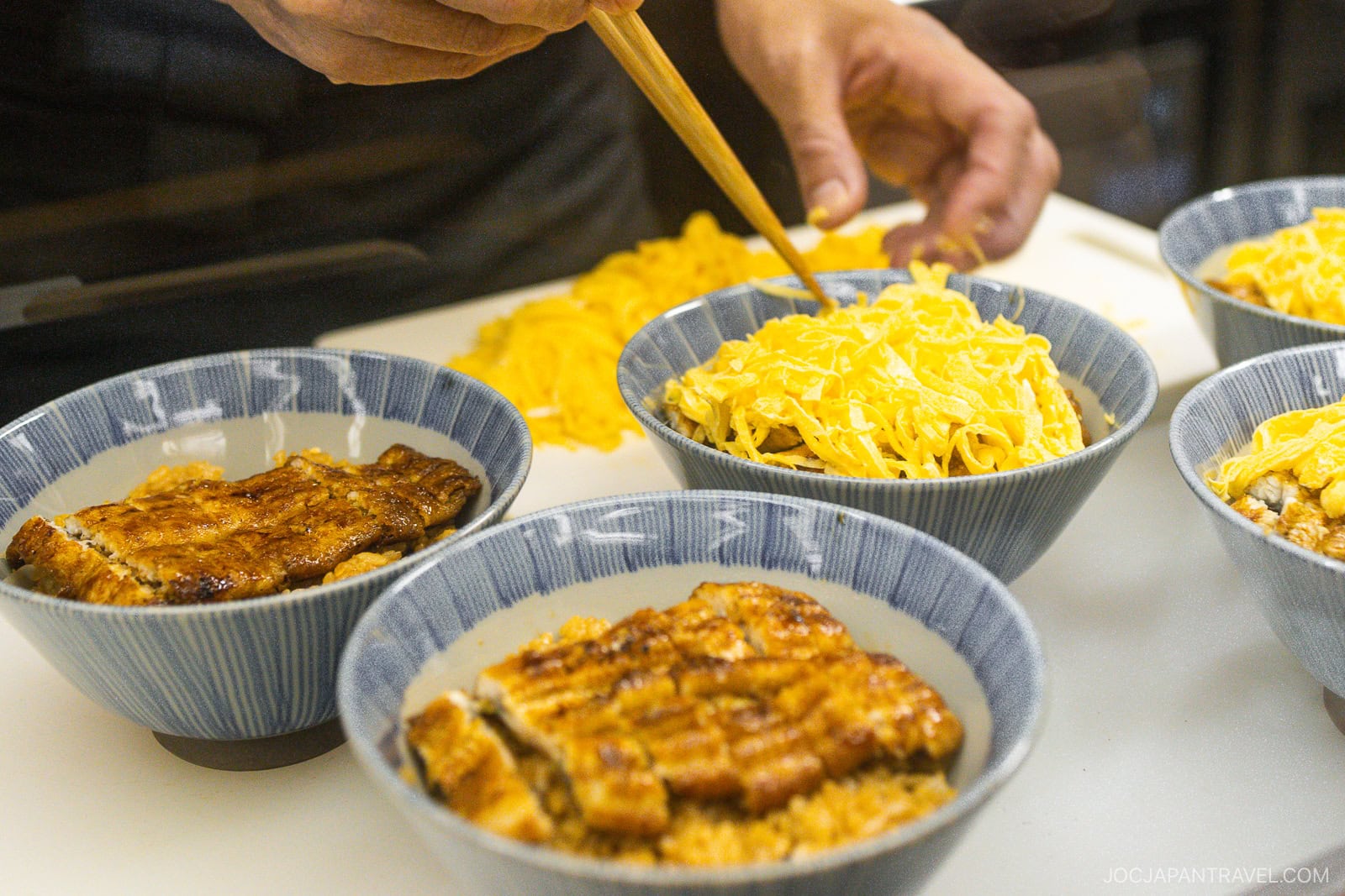 A person uses chopsticks to add shredded egg to bowls of rice topped with grilled eel—a delicious option if you're in Kyoto wondering what to eat, with several prepared bowls arranged on a counter.