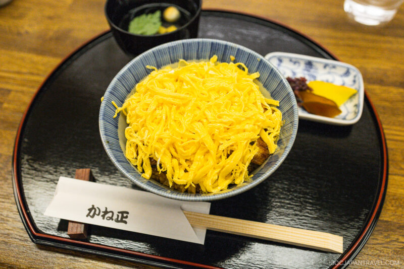 A bowl of Japanese rice topped with shredded egg, served on a black tray with chopsticks, pickles, and a small bowl of soup. If you're in Kyoto wondering what to eat, this traditional set is a delicious local choice.