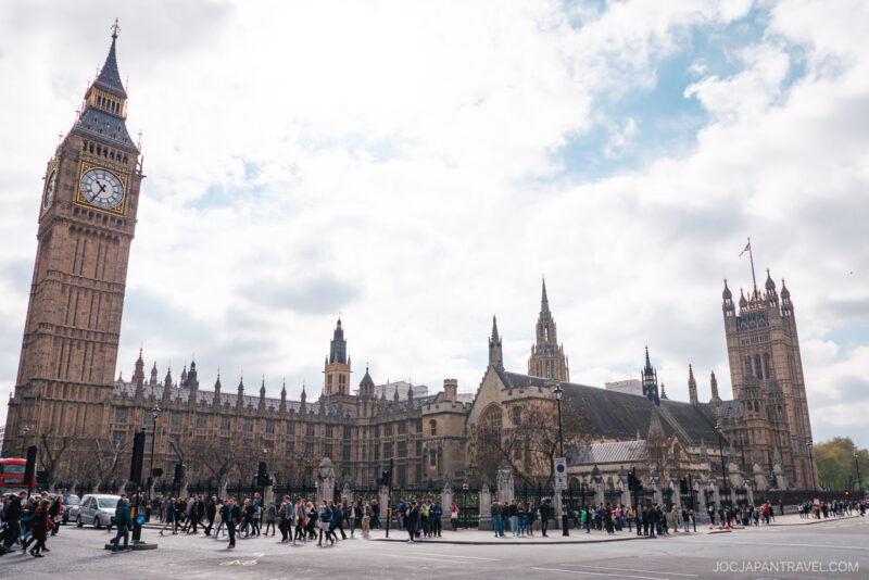 Crowds of people walk in front of the Palace of Westminster and Big Ben in London on a cloudy day, with the iconic clock tower visible on the left and London’s stunning gothic architecture dominating the scene.
