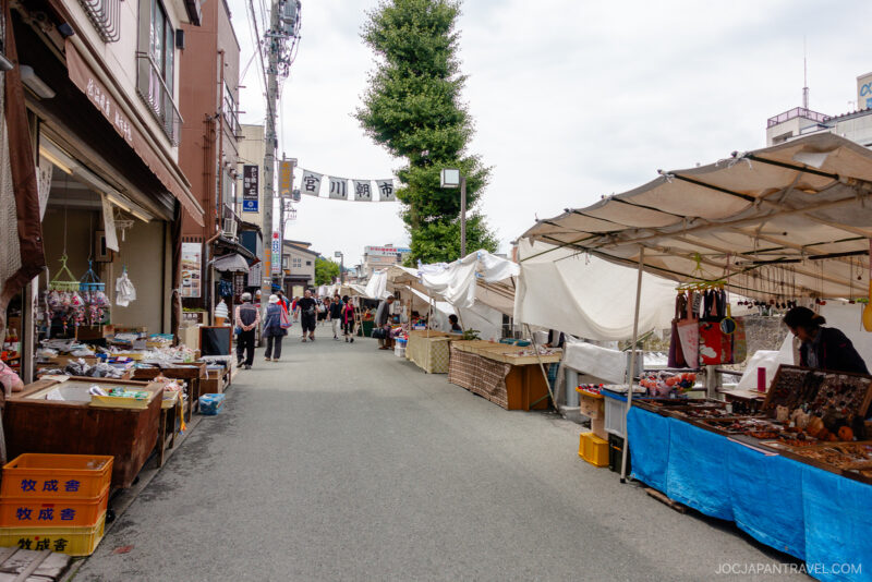 Miyagawa Morning Market is a lively outdoor street in Japan with stalls selling pottery and crafts. Few people stroll the wide pathway, flanked by tents and shops under a partly cloudy sky.