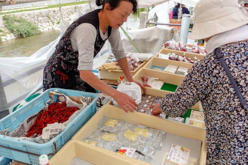 A vendor and a customer interact at an outdoor Miyagawa Morning Market stall selling dried goods, spices, and packaged food, with baskets and trays neatly arranged on the tables.