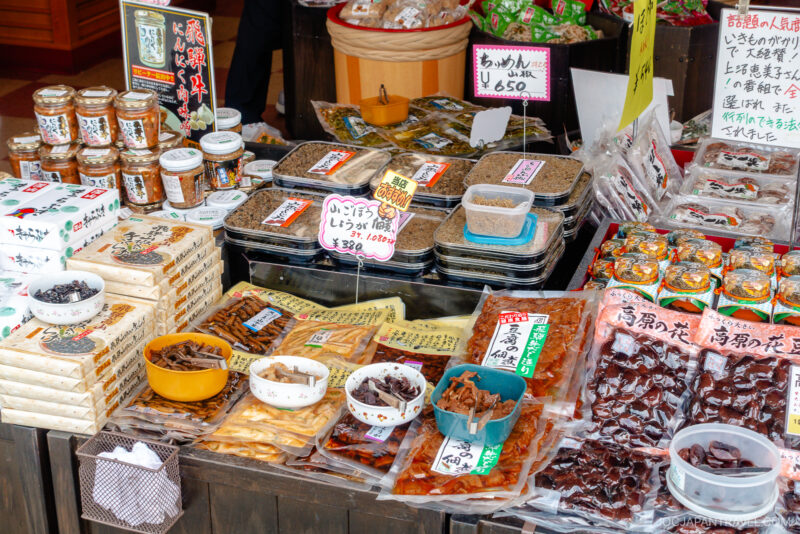 A colorful Miyagawa Morning Market stall displays various packaged Japanese foods, jars, dried goods, and snacks with handwritten Japanese labels and price tags on each item.