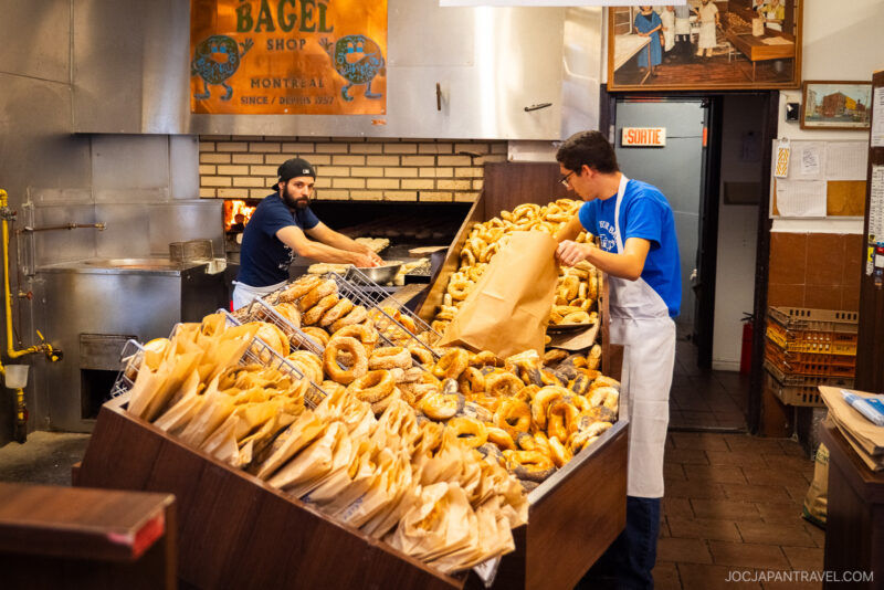 Two workers in a bakery handle large piles of bagels—one fills a paper bag while the other arranges fresh bagels by the brick oven. A sign above reads Bagel Shop Montréal, capturing the spirit of classic Montreal food.