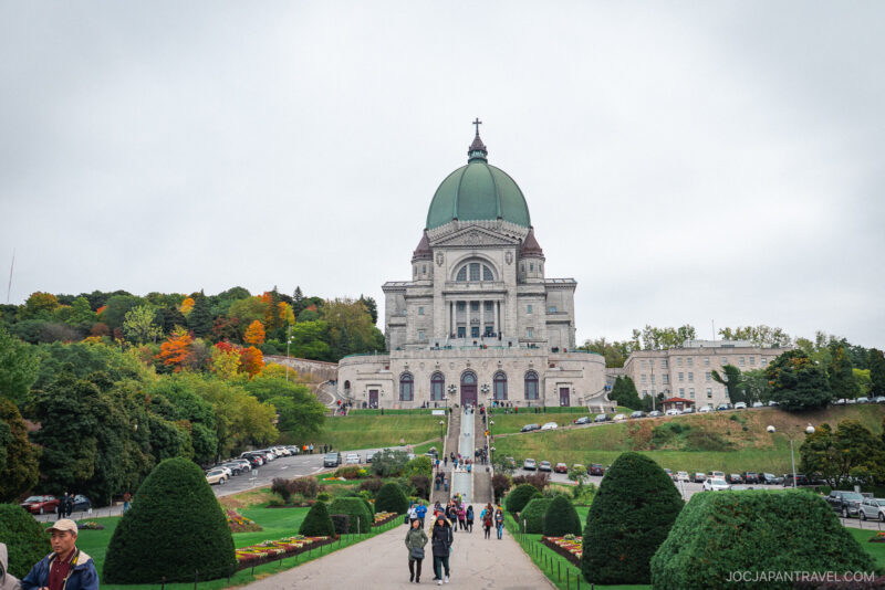 A large stone basilica with a green dome stands atop a hill in Montreal, surrounded by manicured gardens and trees with autumn foliage. People walk up a path toward the entrance, and cars are parked along the roads on either side.