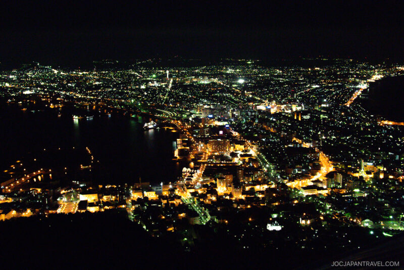 A vibrant cityscape at night with streets and buildings illuminated by countless lights, surrounding a dark bay. The glow of the city extends into the distance under a dark sky, as seen from the summit of Mt. Hakodate.
