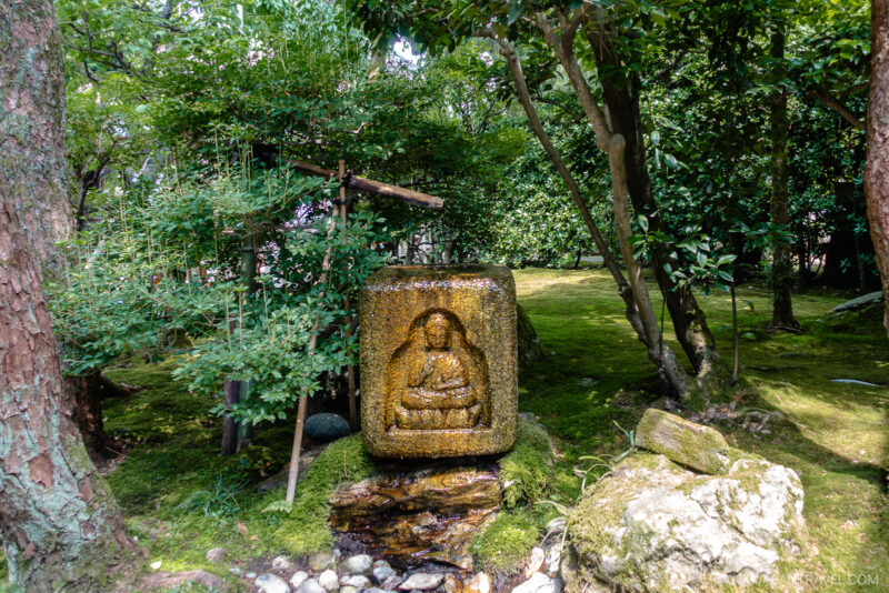A stone relief of a seated Buddha figure stands in the lush, green garden of Myoryu-ji Temple, surrounded by trees and moss-covered ground. Sunlight filters through the foliage, creating a peaceful and serene atmosphere.