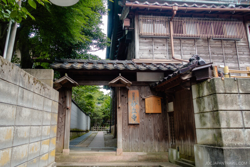 Traditional Japanese wooden temple gate with a tiled roof at Myoryu-ji Temple, surrounded by stone walls and a peaceful, greenery-lined pathway. A wooden sign with Japanese characters hangs beside the entrance.